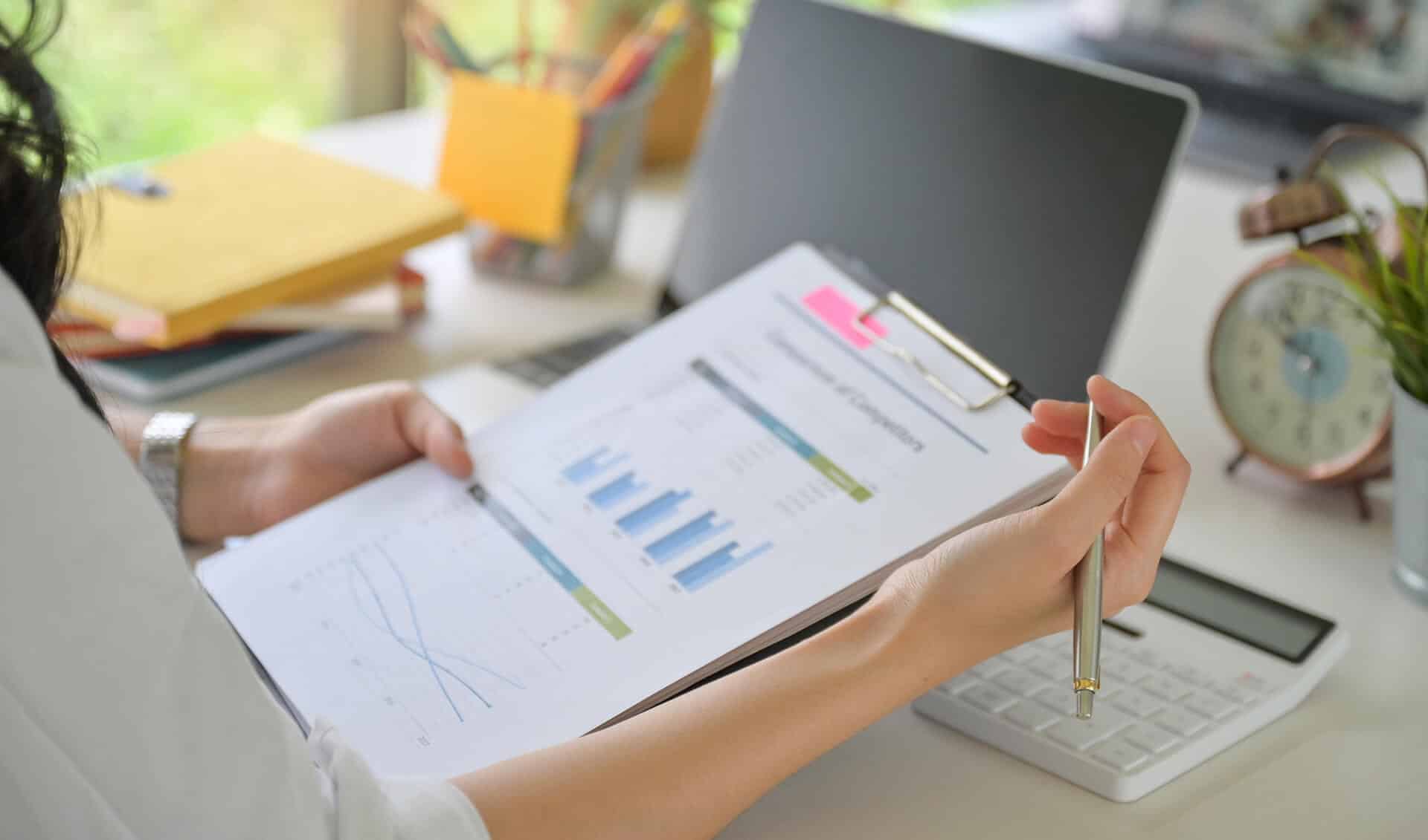 Person reviewing financial charts on a clipboard, holding a pen, with a laptop, calculator, and clock on the desk.