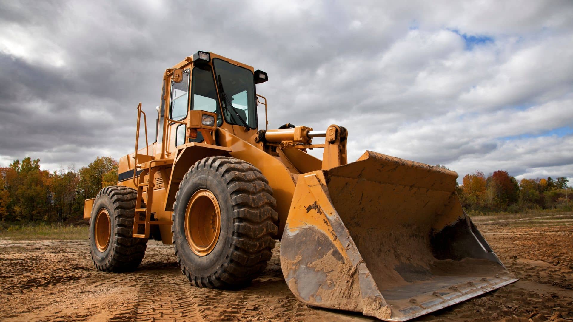 A yellow bulldozer on a dirt field under a cloudy sky, working for the local tree company, is surrounded by trees showcasing vibrant autumn foliage.